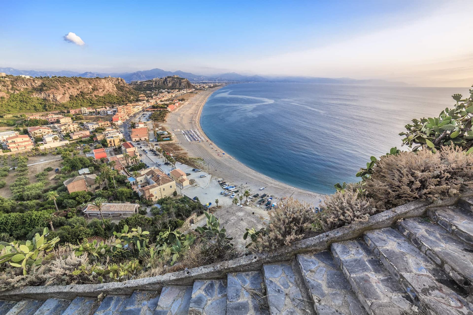 Blick von oben auf Capo Milazzo in Sizilien, Italien, mit einem langen Sandstrand, klarem Wasser und Küstenstadt im Hintergrund