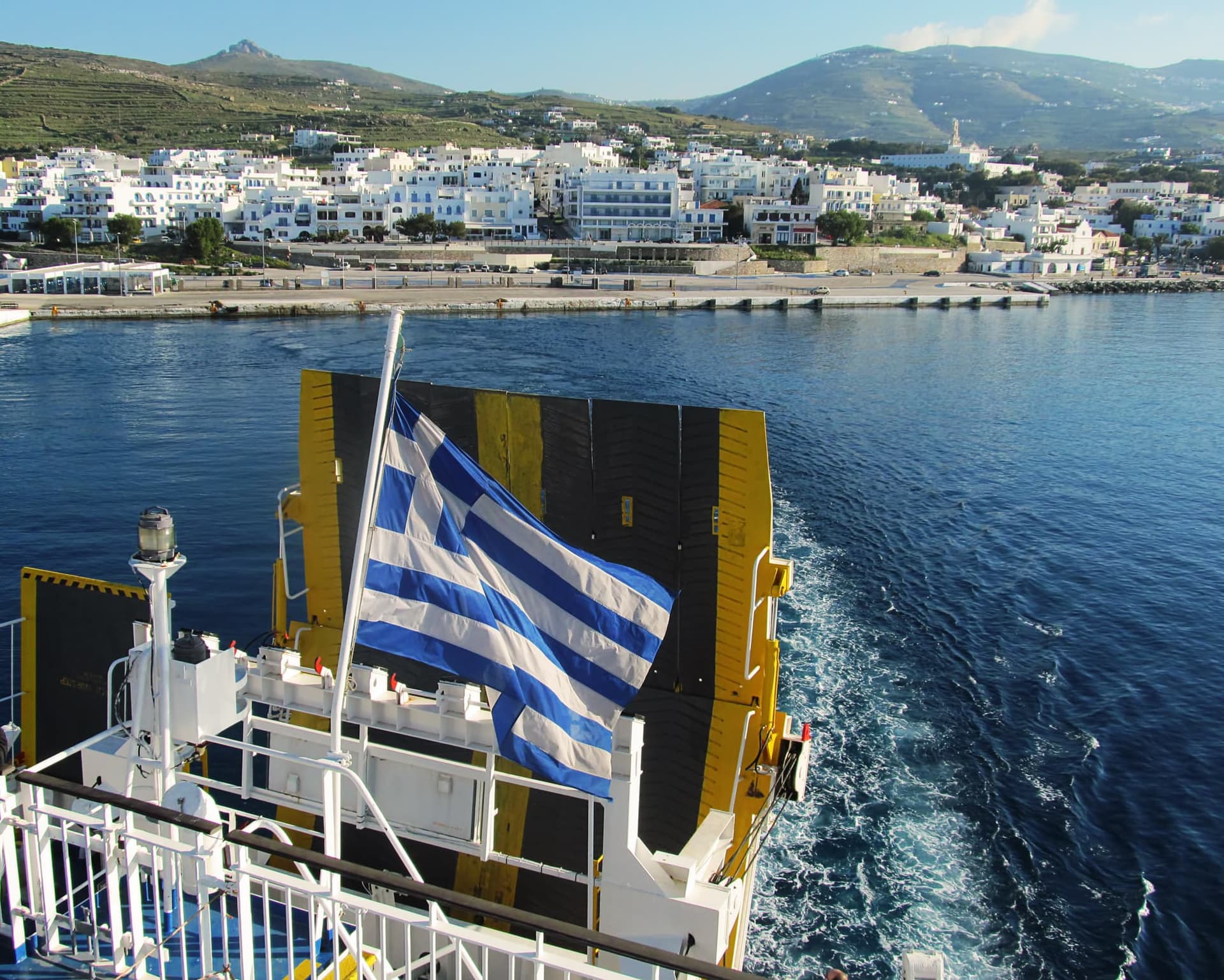 Ferry departing from a Greek island with the Greek flag and a view of the harbor and white houses in the background.