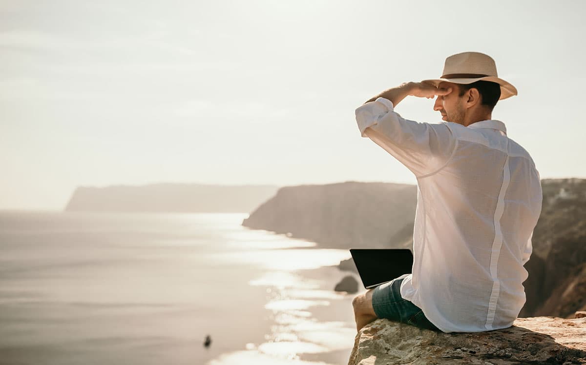 Man sitting on a cliff with a laptop, looking out over the sea near sunset.