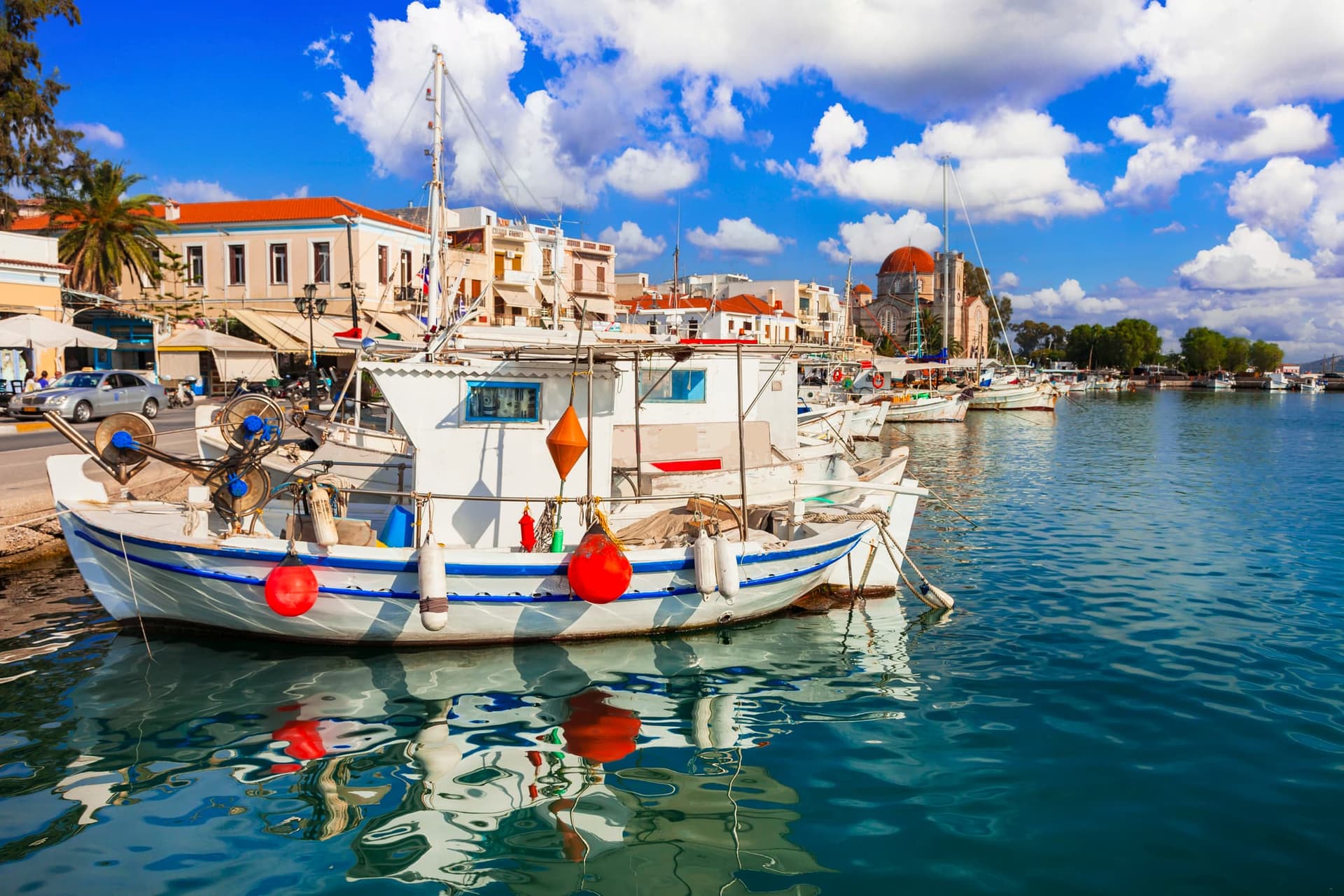 Το γραφικό λιμάνι της Αίγινας, με πολύχρωμα καΐκια, ήρεμα νερά και νεοκλασικά κτίρια δίπλα στη θάλασσα.