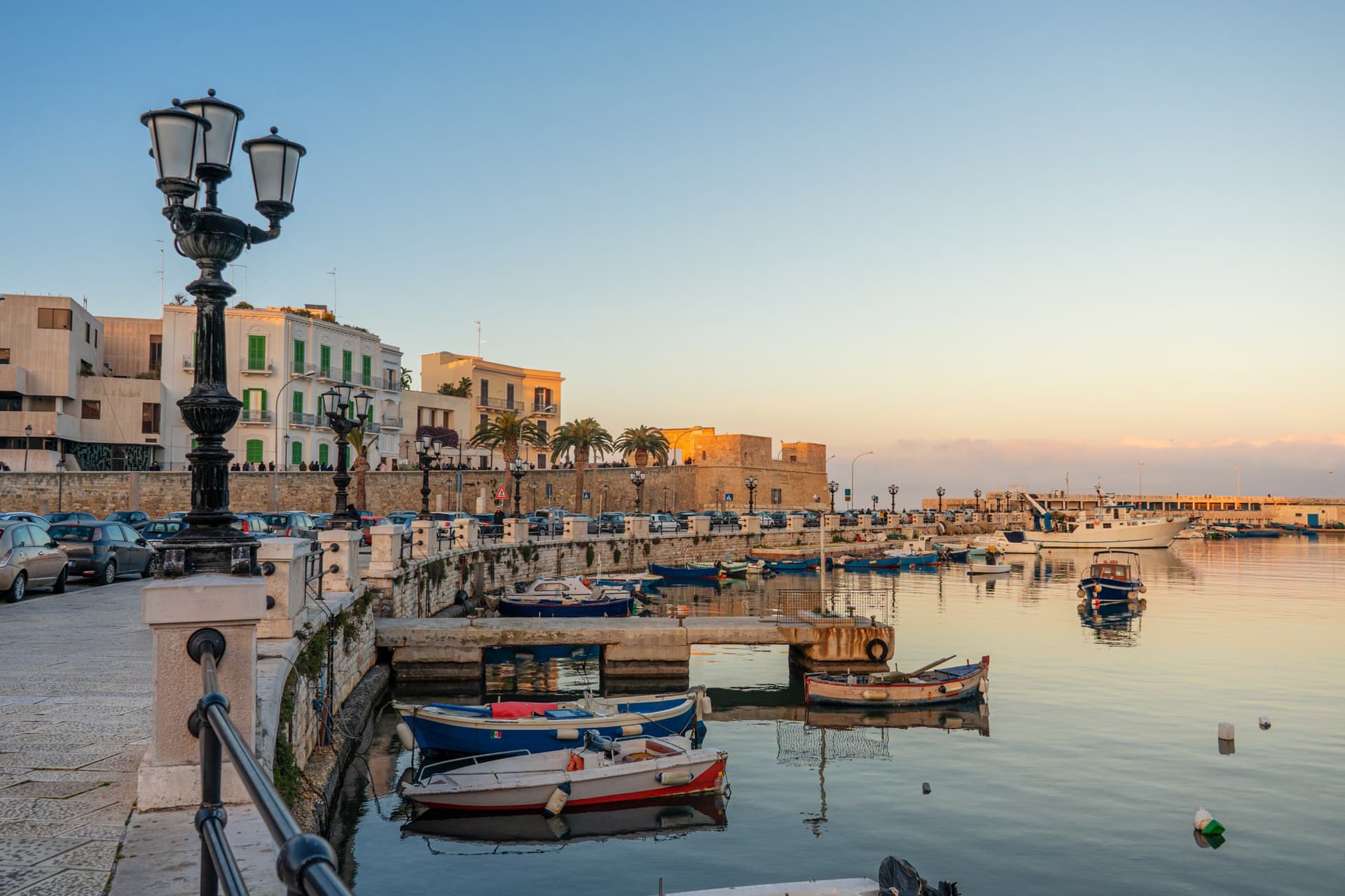 Veduta panoramica di Bari con il suo incantevole porto, piccole imbarcazioni, edifici storici e un lungomare illuminato dal tramonto.