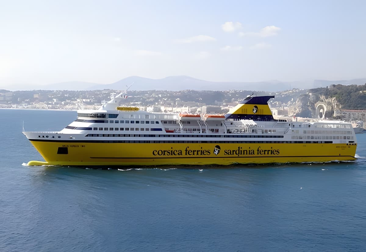 A Corsica Ferries – Sardinia Ferries vessel sailing on calm blue waters, with a coastal city and hills in the background.